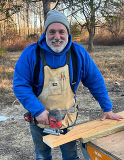 Man in blue jacket and tan apron holding wood plank outdoors