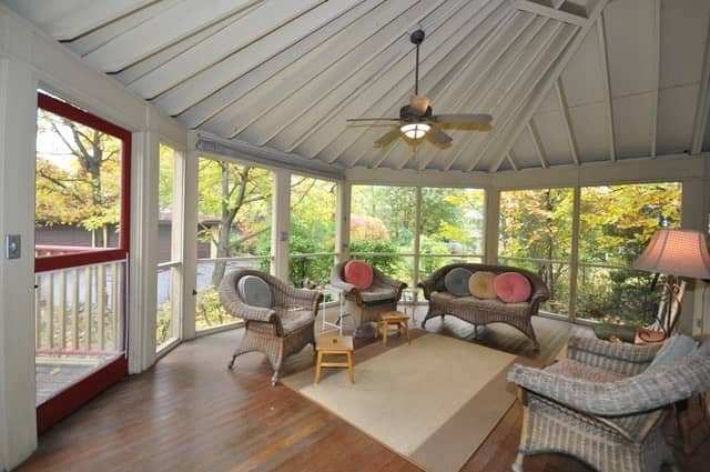 Sunlit screened porch with wicker furniture, ceiling fan, and autumn trees visible through windows